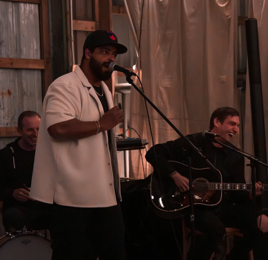 Man singing into a microphone with another person playing guitar in a casual setting.