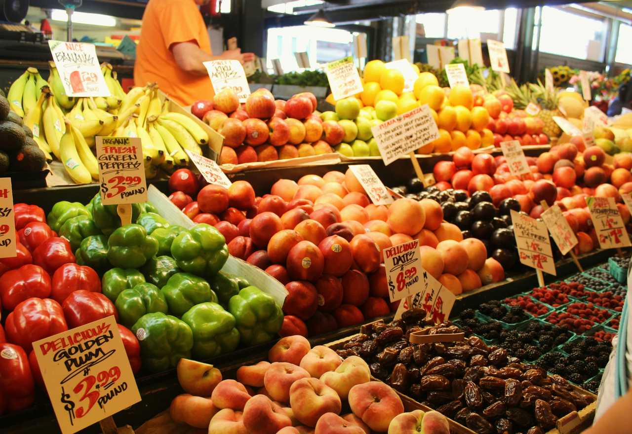 A bustling market scene featuring tiered wooden displays filled with fresh bananas, red apples, peaches, and containers of blackberries. Hand written signs and price tags show the variety of local produce available during the community and retail phase of the farm launch.