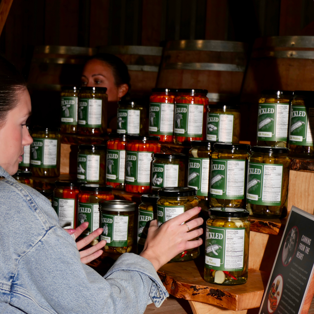 Hands selecting a jar of pickled preserved vegetables from a rustic wooden display at a local market, showcasing handcrafted Farmbox pantry staples.